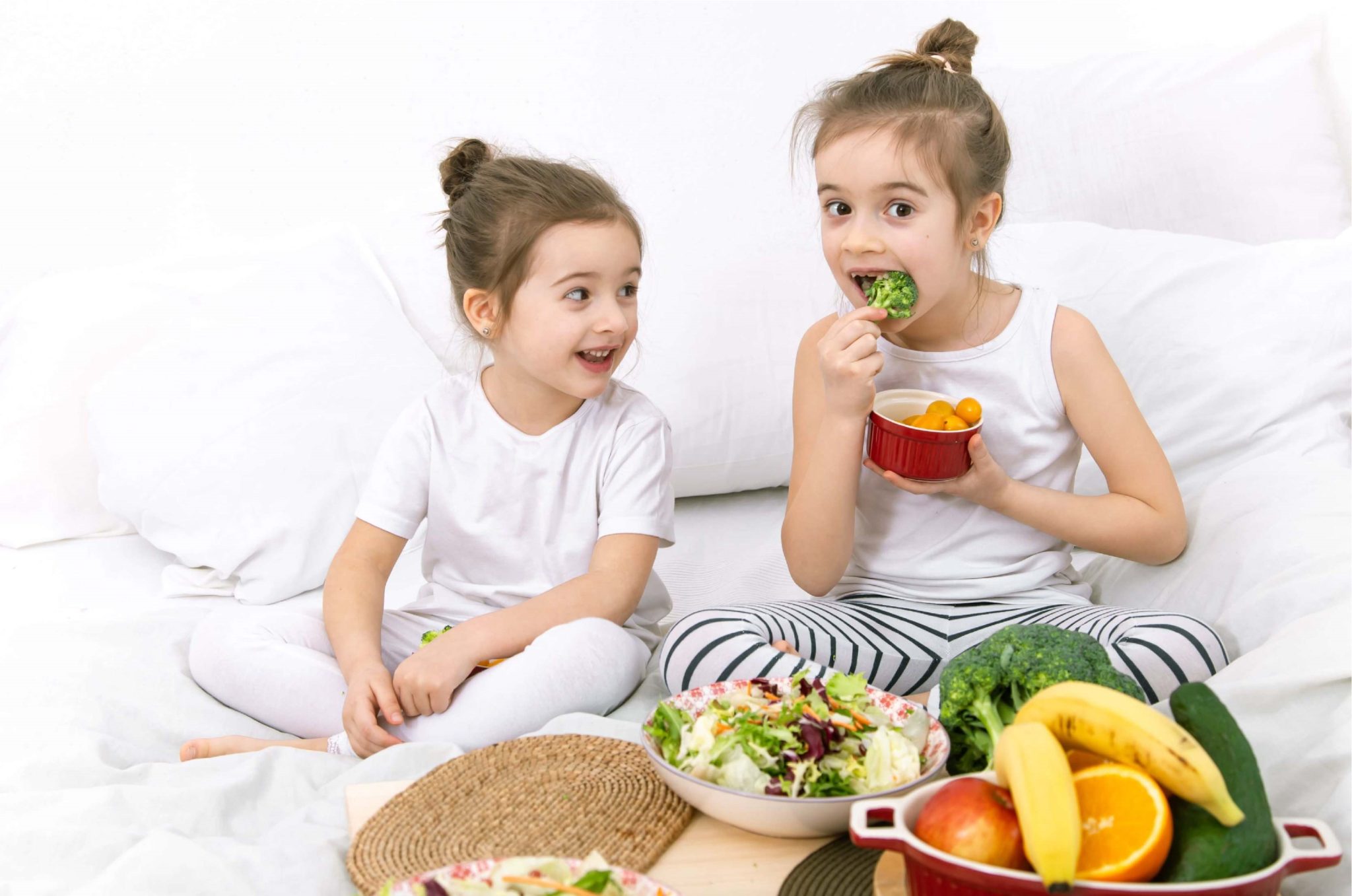 Two children eating broccoli and healthy foods including fruits and vegetables while sitting together.