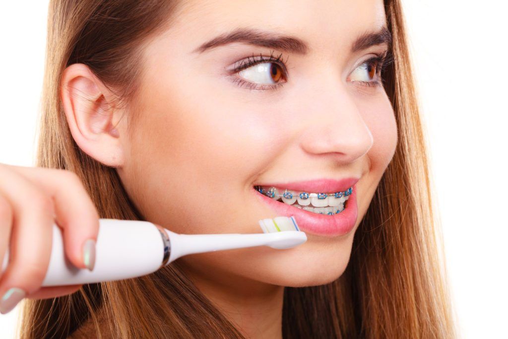 Teenage girl with metal braces smiling while holding an electric toothbrush near her teeth.