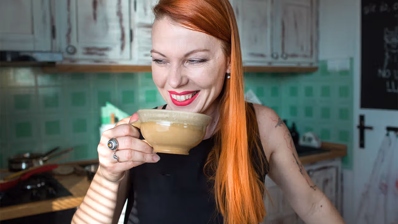 Person smiling while holding and drinking a cup of coffee in a kitchen.